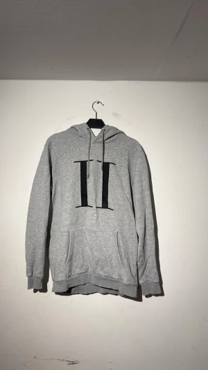 Grå hoodie med svart tryck - Snygg grå hoodie med stor svart tryckt detalj på bröstet. Klassisk känguruficka framtill, justerbar huva med snören och ribbade muddar vid ärmar och nederkant. Perfekt för dig som gillar enkel men stilren streetwear.