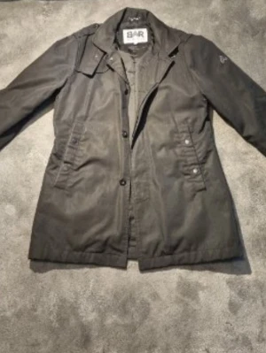 Svart rock från Barbour - Stilren svart rock från Bar Urban med klassisk krage och knäppning framtill. Rocken har flera fickor med lock och knappdetaljer samt en diskret logga på ärmen. Materialet är slitstarkt och har en matt finish, perfekt för en trendig och tidlös look. ✅