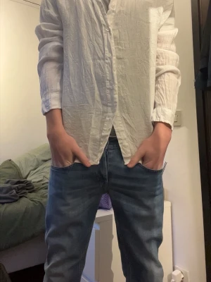 Vit långärmad skjorta i bomull - Klassisk vit skjorta med lång ärm och knappar framtill. Skjortan är tillverkad i ett lätt, skrynkligt bomullstyg som ger en avslappnad vibe. Passformen är normal och skjortan har en enkel, tidlös design utan synliga loggor.