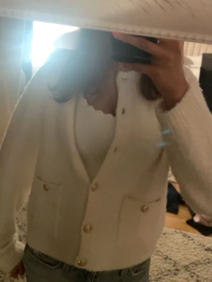 Vit kofta med guldknappar - Mysig vit kofta med v-ringning och dekorativa guldknappar framtill. Koftan har två fickor och är tillverkad i ett fluffigt, stickat material. Perfekt att slänga över en topp för en avslappnad men snygg look.