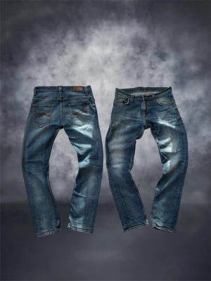 Blå jeans från Nudie Jeans, W32 L32 - Säljer ett par klassiska blå jeans från Nudie Jeans i storlek W32 L32. Jeansen har en snygg tvätt med slitningar, raka ben och fem fickor. Tillverkade i slitstarkt denim med orange sömmar och läderpatch bak i midjan. Perfekta för en avslappnad och trendig look.