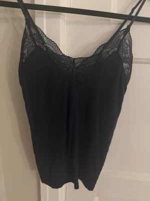 Mörkblå spetslinnetopp från Lindex  - Snygg mörkblå linnetopp från Lindex med tunna axelband och dekorativ spets längs urringningen. Tillverkad i en mjuk Tencel lyocell-blandning som känns skön mot huden. Perfekt att styla lager på lager eller bära som den är.