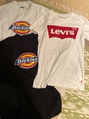 T-shirtpaket Dickies & Levi's logga - Säljer tre snygga t-shirts: två från Dickies (en svart och en vit) med stor färgglad logga på bröstet, samt en vit Levi's t-shirt med klassisk röd logga. Alla har rund hals och korta ärmar. Perfekta till jeans eller shorts för en avslappnad streetstil. Det kan förekomma skador.