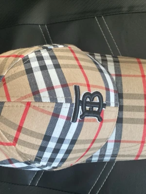 Beige keps med rutigt mönster - Snygg keps i klassiskt beige rutigt mönster med svarta, vita och röda linjer. Kepsen har en böjd skärm och broderad svart logotyp framtill. Justerbar baktill och tillverkad i ett lätt tygmaterial som andas.