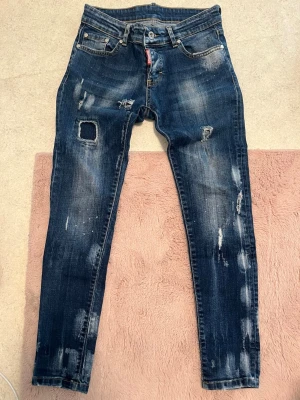 Mörkblå jeans från Dsquared2 - Säljer ett par mörkblå jeans från Dsquared2 med coola slitningar, hål och patch-detaljer. Jeansen har smal passform och klassisk femficksdesign. Perfekta för dig som gillar en edgy streetstil och vill sticka ut med snygga detaljer.