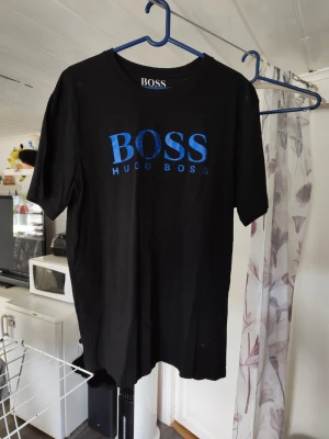 Svart Hugo Boss t-shirt med blå logga - Snygg svart t-shirt från Hugo Boss med stor blå logga på bröstet. Klassisk rund halsringning och korta ärmar. Perfekt för dig som gillar stilrena och trendiga plagg med tydlig branding. Mer som xl än xxl 