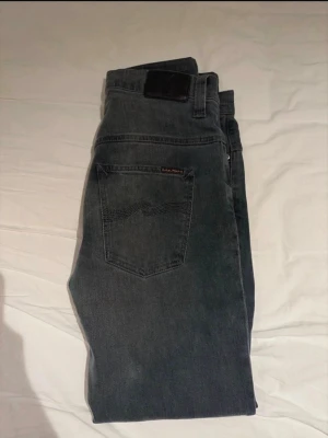 Mörkgråa jeans från Nudie Jeans Tape Ted - Säljer ett par mörkgråa jeans från Nudie Jeans med klassisk femficksmodell och diskret logga på bakfickan. Jeansen har en rak passform och är tillverkade i mjuk denim. Perfekta för dig som gillar en clean och stilren look.Måtten står i bilderna. Storlek W30 L32. Fraktas samma dag. Hör av dig vid intresse!