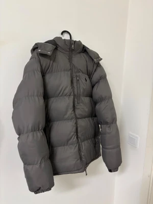 Mörkgrå dunjacka med huva - Säljer en mörkgrå dunjacka med avtagbar huva och dragkedja framtill. Jackan har en bröstficka med dragkedja och två sidofickor. Puffig modell som håller dig varm under kalla dagar. Perfekt för vintern och riktigt snygg till streetwear-stilen. Modellen är L men det står 2 xl pga kinesiska sizes