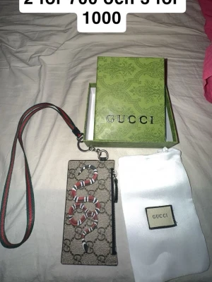 Gucci plånbok med orm och axelrem - Snygg plånbok från Gucci med klassiskt GG-mönster och en röd-vit orm på framsidan. Plånboken har en avtagbar axelrem i grönt och rött samt dragkedja. Kommer med original ask och dustbag. Perfekt accessoar för dig som gillar lyxiga detaljer.