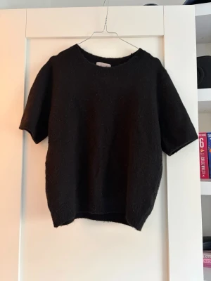Svart stickad tröja med kort ärm - En enkel och stilren svart stickad tröja med korta ärmar och rund hals. Tröjan har en rak passform och är gjord i ett mjukt material som känns skönt mot huden. Perfekt att matcha med jeans eller kjol för en avslappnad look.