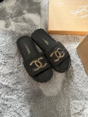 Svarta Chanel tofflor med guldlogo - Säljer ett par svarta tofflor från Chanel med ikonisk guldig CC-logo framtill. Tofflorna har en grov textur och öppen tå, perfekta för en lyxig och chill stil. Passar dig som vill ha något både trendigt och klassiskt.