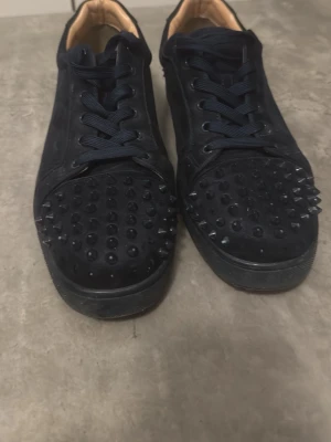  Louboutin sneakers med nitar - Säljer ett par svarta sneakers från Christian Louboutin med coola svarta nitar på tån och klassisk röd sula. Skorna har lågt skaft, svarta skosnören och är tillverkade i mocka. Perfekta för dig som vill sticka ut med en edgy stil. Det har lossnat några spikar där med det låga pris