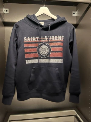 Mörkblå Saint Laurent hoodie - Mörkblå hoodie från Saint Laurent med tryck i vitt och rött framtill. Stor ficka på magen och justerbar huva med snören. Trycket har en vintage-look med texten 'SAINT LAURENT' och SL-emblem i mitten. Perfekt för en avslappnad och cool stil. Skick är 10/10 de är stolek S men tycker den passar Xs! 