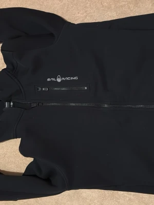 Svart hoodie från Sail Racing - Svart hoodie från Sail Racing med dragkedja framtill och en extra bröstficka med dragkedja. Hoodien har justerbar huva med snörning och logga på bröstet samt på huvan. Perfekt för dig som gillar stilren och sportig design.