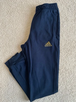 Mörkblå Real Madrid mjukisbyxor Adidas - Mörkblå mjukisbyxor från Adidas med Real Madrid-logga och Adidas-logga i guld på framsidan. Byxorna har resår i midjan och vid bensluten samt sidofickor. Perfekta för chill eller träning.