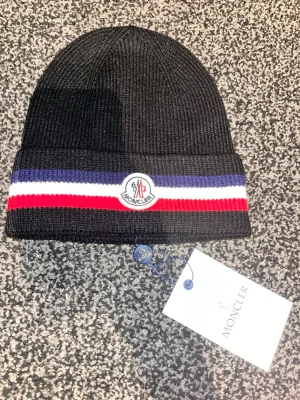 Svart Moncler mössa med ränder - Svart ribbstickad mössa från Moncler med bred uppvikt kant och klassisk logotyp framtill. Mössan har dekorativa ränder i blått, vitt och rött runt kanten. Perfekt accessoar för att hålla värmen och ge en sportig touch till din stil.