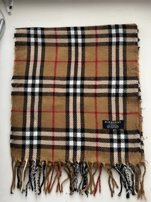 Beige rutig halsduk från Burberry - Klassisk beige halsduk från Burberry i 100% kashmir med det ikoniska rutmönstret i svart, vitt och rött. Halsduken har fransar i ändarna och är tillverkad i England. Perfekt accessoar för att lyfta din outfit.