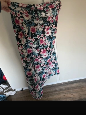 Blommig långkjol med tropiskt mönster - Långkjol med färgstarkt blommönster i rosa, vitt och grönt på mörk botten. Kjolen har rak passform och är tillverkad i ett mjukt, lätt material som passar perfekt till varmare dagar. Ingen synlig dragkedja eller knappar. Man knyter kjolen, kan även knytas om till en topp med rätt teknik.