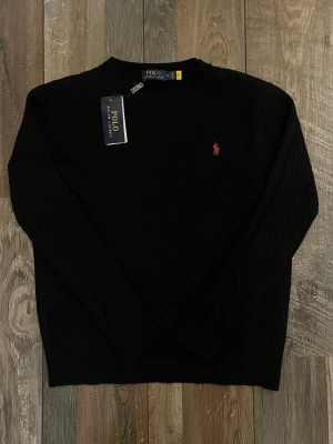 Svart kabelstickad tröja Polo Ralph Lauren - Kabelstickad svart tröja från Polo Ralph Lauren med rund halsringning och röd broderad logga på bröstet. Är lite liten i storlek passar som S och är slimfit.