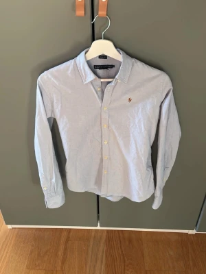 Ljusblå skjorta från Ralph Lauren - Snygg ljusblå skjorta från Ralph Lauren. Den är knappt använd och därav i bra skick. Hör av dig vid frågor och funderingar.Priset är ej hugget i sten. Skjortan är en kvinnlig version men går att användas utav killar också om man har en bredare midja.