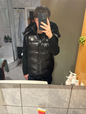 Svart dunväst från Moncler med huva - Snygg svart dunväst från Moncler med glansig finish och avtagbar huva. Västen har två dragkedjefickor framtill och klassisk Moncler-logga på bröstet. Perfekt för lager-på-lager och streetstyle. Sitter bra på mig som är 170