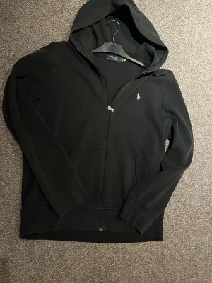 Svart hoodie från Polo Ralph Lauren - Svart Ralph Lauren zip-up hoodie. Klassisk broderad logga på bröstet. Skön och stilren modell som passar perfekt till jeans. Funkar till de flesta stilarna. Ända dåliga är att den saknar snöre men annars inga fel. Säljer för den är för stor (Köp eller byta mot en likadan i storlek S)