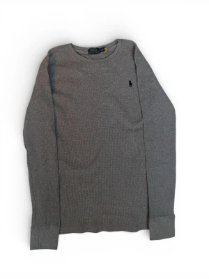 Grå stickad tröja från Polo Ralph Lauren - Snygg grå stickad tröja från Polo Ralph Lauren med klassisk rund hals och lång ärm. Tröjan har diskret svart broderad logga på bröstet och är tillverkad i ett mjukt, strukturerat material. Perfekt för lager-på-lager och enkel att matcha.