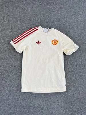 Vit Manchester United t-shirt Adidas - Vit t-shirt från Adidas med Manchester United-logga på bröstet och klassiska röda ränder längs axlarna. T-shirten har rund hals och korta ärmar, perfekt för dig som gillar sportig stil och fotboll.