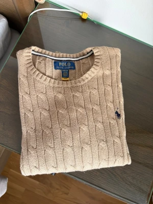 Beige kabelstickad tröja Polo Ralph Lauren - Kabelstickad beige tröja från Polo Ralph Lauren med rund halsringning och broderad marinblå logga på bröstet. Klassisk och tidlös design i mjukt material, perfekt för lager-på-lager-stil.