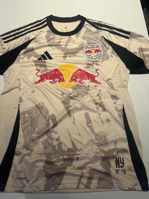 New York Red Bulls matchtröja Adidas - Snygg beige och svart matchtröja från Adidas för New York Red Bulls med klubbmärke och Red Bull-logga på bröstet. Tröjan har digitalt camo-mönster, svarta detaljer och klassiska Adidas-ränder på axlarna. Tillverkad i lätt och ventilerande material.