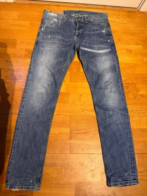 Blå raka dondups  - Säljer ett par blå jeans med raka ben och snygga slitningar samt ljusa tvättade partier. Jeansen har fem fickor och klassisk knappgylf. Materialet är denim i bomull och de har en avslappnad, trendig look.