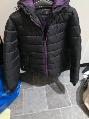 Svart pufferjacka från High Mountain - Säljer en svart pufferjacka från High Mountain med lila detaljer och dragkedja. Jackan har huva och är vadderad för extra värme. Insidan är fodrad i lila och modellen är midjelång med två fickor framtill. Perfekt för kalla dagar!