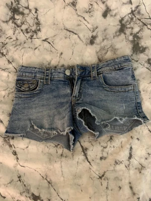Blå jeansshorts med råa kanter - Snygga blå jeansshorts med råa, fransiga kanter och slitningar. Klassisk femficksmodell med knapp och dragkedja framtill samt fickor med lock bak. Perfekta för sommaren och ger en avslappnad vibe.