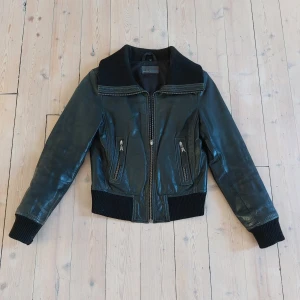 Svart skinnjacka från ROCK'NBLUE - Snygg svart skinnjacka ROCK'NBLUE med bred krage och ribbade muddar vid ärmslut och midja. Jackan har dragkedja framtill och två snedställda fickor med dragkedjor. Perfekt för dig som vill ha en cool och tidlös look. Kommer från ett djur och rökfritt hem. 