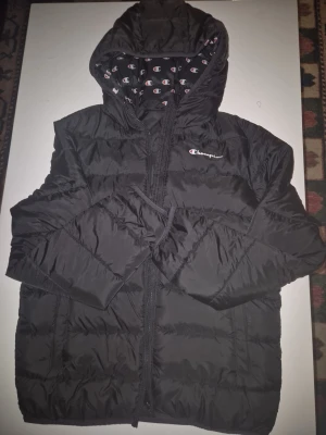 Svart Champion pufferjacka med huva - Svart pufferjacka från Champion med huva och dragkedja. Jackan har ett mönstrat innerfoder med Champion-loggor och broderad logga på bröstet. Tillverkad i lätt, vadderat material som håller värmen under kalla vinterdagar. Klassisk och sportig design.