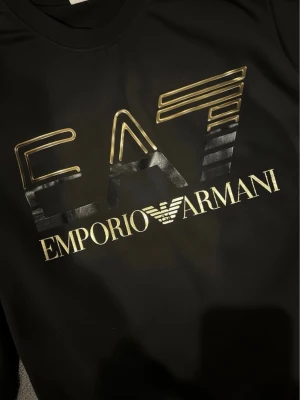 Svart EA7 Emporio Armani t-shirt - Svart t-shirt från EA7 Emporio Armani med stor, glansig logga i guld och svart framtill. Klassisk rund halsringning och korta ärmar. Perfekt för dig som gillar exklusiva streetwear-plagg med tydlig logotyp.
