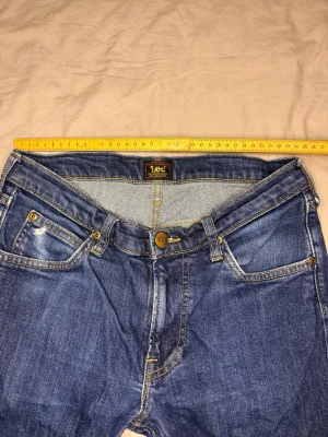 Blå jeans från Lee med raka ben - Snygga blå jeans från Lee med klassisk femficksdesign och raka ben. Jeansen har guldfärgade nitar och kontrastsömmar, samt Lee-logga på knappen och bak i linningen. Perfekta för en avslappnad och stilren look.