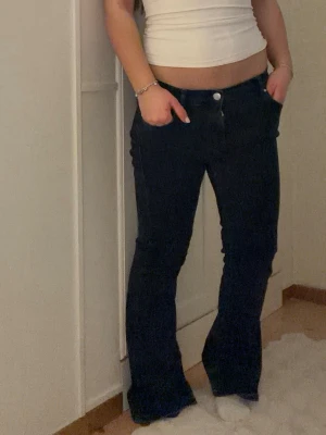 Mörkblå bootcut jeans med låg midja - Säljer ett par mörkblå jeans med bootcut-modell och låg midja. Jeansen har klassisk femficksdesign och silverfärgade knappar. Snygga att styla med crop top eller hoodie för en trendig look.
