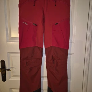 Röda vandringsbyxor från Lundhags - Pigga, smidiga,  röda vandringsbyxor från Lundhags med förstärkta partier i mörkrött och svarta detaljer vid bensluten. Byxorna har flera dragkedjeförsedda fickor och är tillverkade i slitstarkt material. Kardborre vid benslut. Bra skick.