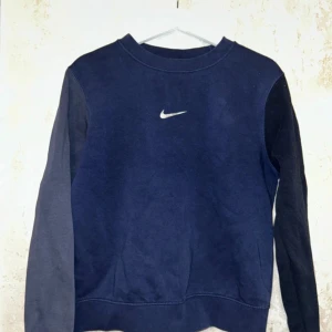Mörkblå Nike sweatshirt  - Säljer en mörkblå crewneck sweatshirt från Nike i storlek XS. Tröjan har klassisk Nike-logga på bröstet och är tillverkad i mjuk bomullsblandning. Ärmarna är i olika nyanser av blått för en cool kontrast. Perfekt för chill dagar eller till skolan.