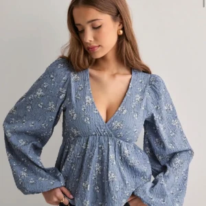 Blommig blå blus med stora ärmar - Supersöt blå blus med blommigt mönster och v-ringad omlottfront. Vida långa ärmar. Blusen har aldrig kommit till användning och är därför som ny, den är i storlek 34 alltså xs men den är i stretchigt material💗