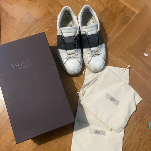 Valentino Open sneakers - Snygga Valentino Garavani Open sneakers i vitt skinn med bred svart rem över vristen. Klassisk rund tå, platt sula och diskreta nitar baktill. Kommer med originalkartong och dustbags. Perfekta för dig som gillar exklusiva och trendiga sneakers.