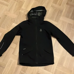 Svart vindjacka från Haglöfs med huva - Svart vindjacka från Haglöfs med minimalistisk design, hel dragkedja och fast huva. Jackan har diskreta reflexdetaljer på ärmen och två snedställda fickor framtill. Perfekt för blåsiga dagar och outdoor-aktiviteter.