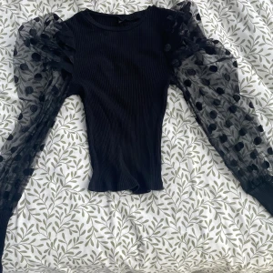 Svart ribbad topp med puffärmar i mesh - Supersnygg svart topp med ribbad kropp och transparenta puffärmar i mesh med stora svarta prickar. Ärmarna är långa och avslutas med breda ribbade muddar. Perfekt för dig som vill sticka ut med coola detaljer och trendig look.
