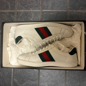 Gucci Ace vita sneakers med gröna detaljer - Gucci Ace sneakers i vitt skinn med klassiska gröna och röda ränder på sidan. Baksidan har en grön krokopräglad detalj med Gucci-logga. Platt sula, snörning och rund tå. Perfekta för dig som vill ha en clean men lyxig look.