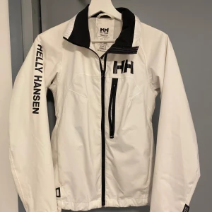 Vit vindjacka från Helly Hansen - Snygg vit vindjacka från Helly Hansen med svart HH-logga på bröstet och tryck på ärmen. Jackan har hög krage, svart dragkedja, bröstficka med dragkedja och meshfoder på insidan. Perfekt för blåsiga dagar och sportig stil.