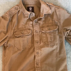Beige overshirt från Brandit S - Säljer en beige overshirt från Brandit i storlek S. Skjortan har två bröstfickor med lock och knappar, axelklaffar och klassisk krage. Tillverkad i bomull och perfekt för en avslappnad streetstil.