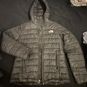 Svart pufferjacka från The North Face - Säljer en svart pufferjacka från The North Face i storlek M. Jackan har huva, två dragkedjeförsedda fickor och quiltad design. Perfekt för kyliga dagar och har en stilren look med logga på bröstet. Materialet är syntet och jackan stängs med dragkedja.