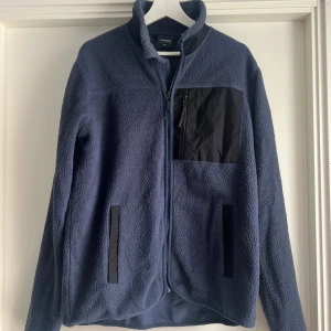 Blå fleecejacka från Everest M - Mysig blå fleecejacka från Everest med svarta detaljer och dragkedja framtill. Jackan har en svart bröstficka med dragkedja och två sidofickor. Perfekt för kyliga dagar och outdoor-häng. Broderad logga på ärmen och hög krage för extra värme.