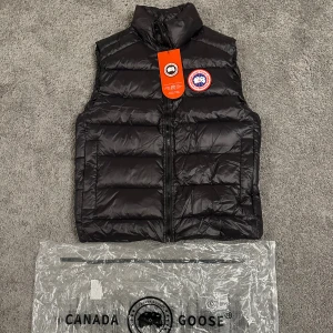 Svart dunväst från Canada Goose - Snygg svart dunväst från Canada Goose med klassisk logga på bröstet. Västen har quiltad design, hög krage och dragkedja framtill. Perfekt att slänga över en hoodie för extra värme. Kommer med originalpåse från Canada Goose.